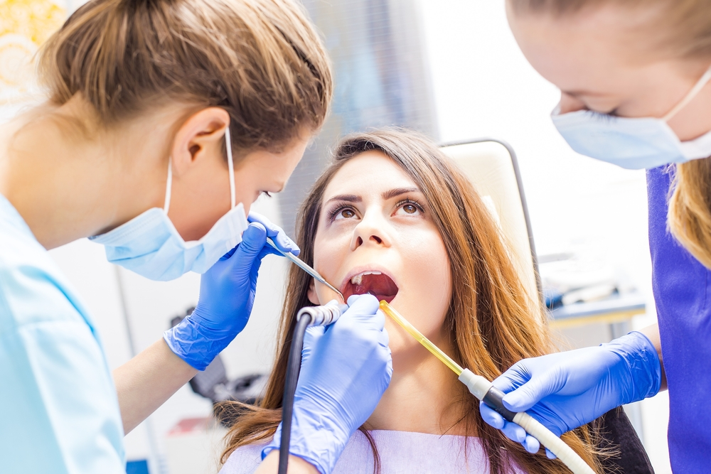 A woman sits in a dental chair while a dentist examines her teeth with a dental tool.